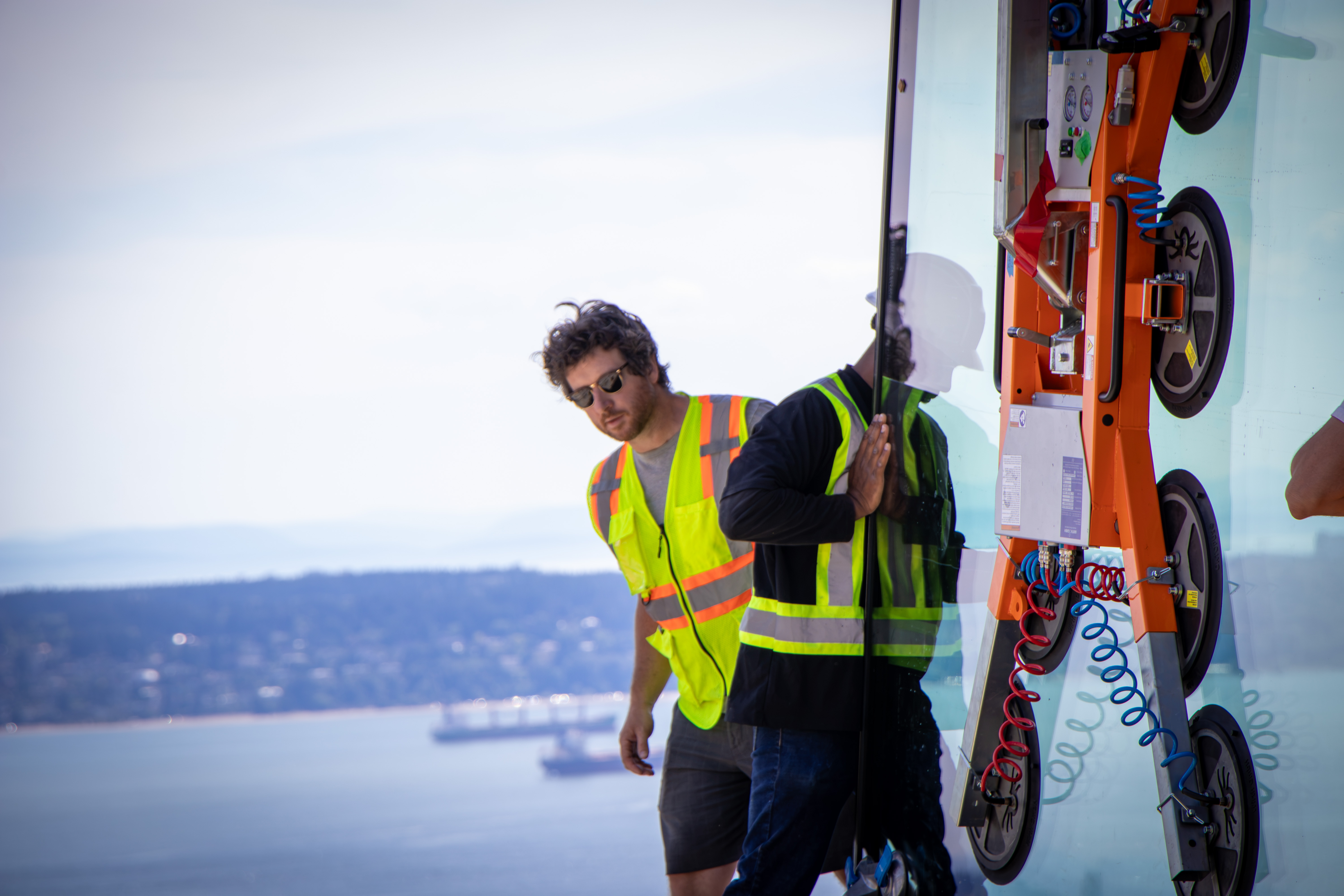 Glazing installers in hard hats and high-visibility vests guide a large glass panel with a vacuum lifter at a construction site overlooking water and a distant city skyline.